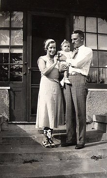 A couple stand on the front steps of a house. The man, aged about 30, dressed in grey flannels and a white shirt, holds a baby girl in his arms and gazes at her. The woman, dressed in a frock and co-respondent shoes from the 1930s stands next to them, touching the baby girl and smiling at the câmera. The baby is dressed in a white frilly frock, white shoes and with a white ribbon in her hair.