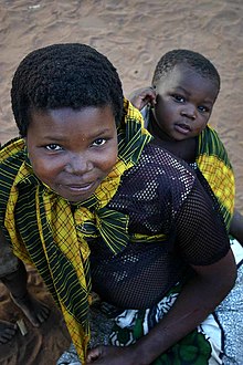 A young mother smiles up at the câmera. On her back is her baby gazing at the câmera with an expression of lively interest.